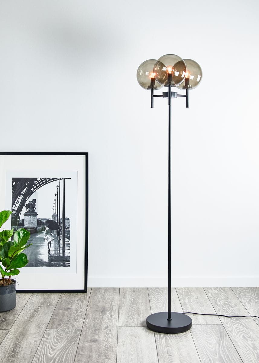 Crown lampa podłogowa 3-punktowa w kolorze czarnym z przydymionymi kloszami. Stojąca, z długim metalowym trzonkiem i okrągłą podstawą. W tle widoczna roślina w doniczce oraz czarno-biała fotografia na ścianie.