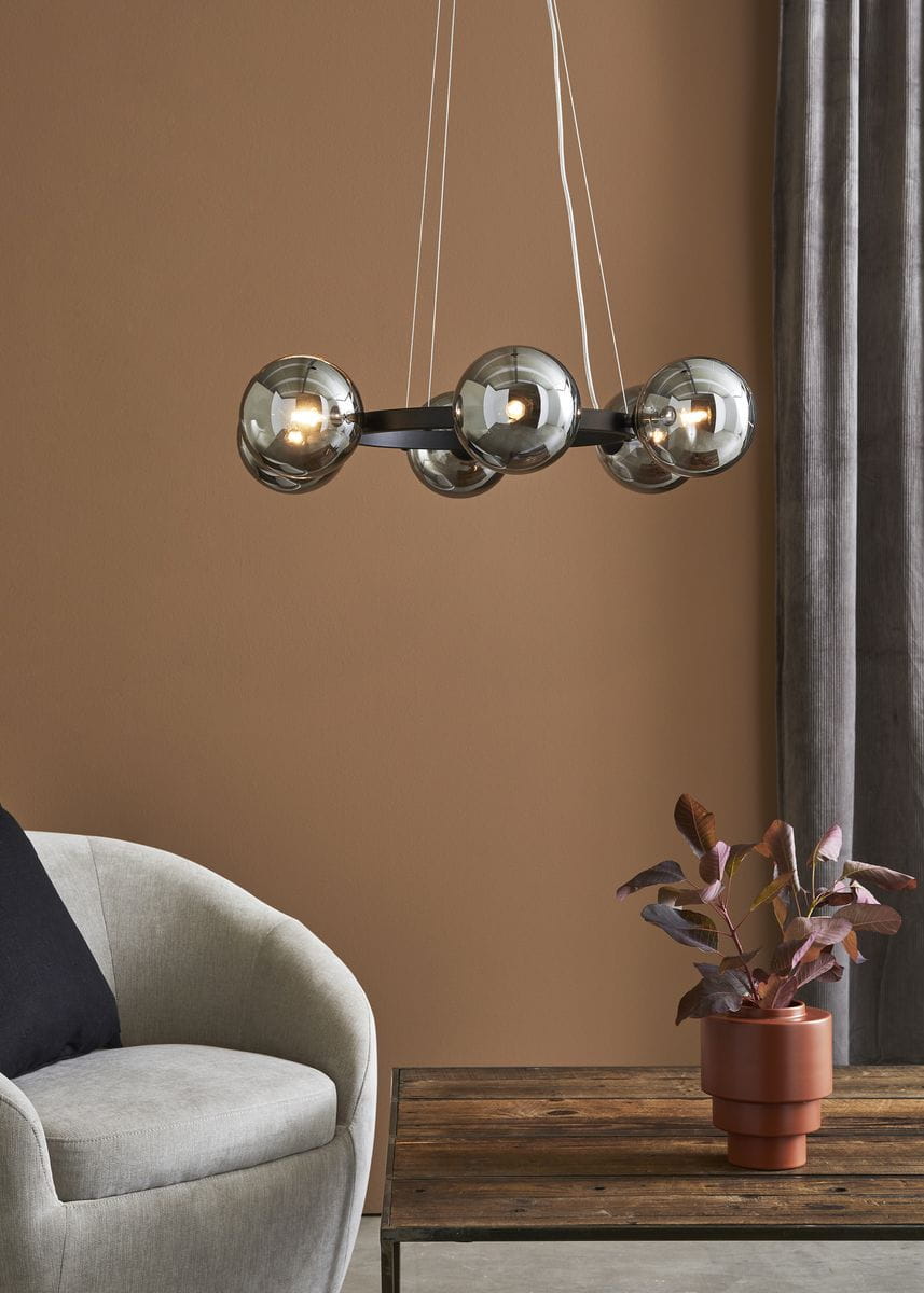 Lampa wisząca Circle 6-punktowa, czarna z dymionymi kloszami, zawieszona na cienkich linkach w minimalistycznym stylu. Widoczna w aranżacji z szarym fotelem i drewnianym stolikiem, w tle ściana w odcieniu brązu.