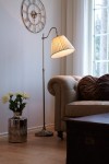 Lampa podłogowa Charleston w kolorze patyny z beżowym kloszem, ustawiona obok sofy. Stojak o regulowanej wysokości, z okrągłą podstawą. W tle widoczny zegar ścienny oraz kwiaty w metalowym wazonie.