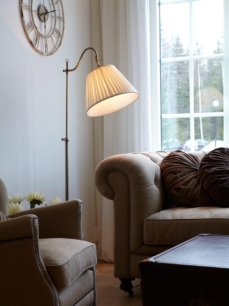 Lampa podłogowa Charleston 1-punktowa w kolorze patynowym z beżowym kloszem, o plisowanej fakturze, ustawiona obok wygodnego fotela w nowoczesnym wnętrzu. W tle widoczny zegar ścienny i okno z firankami.