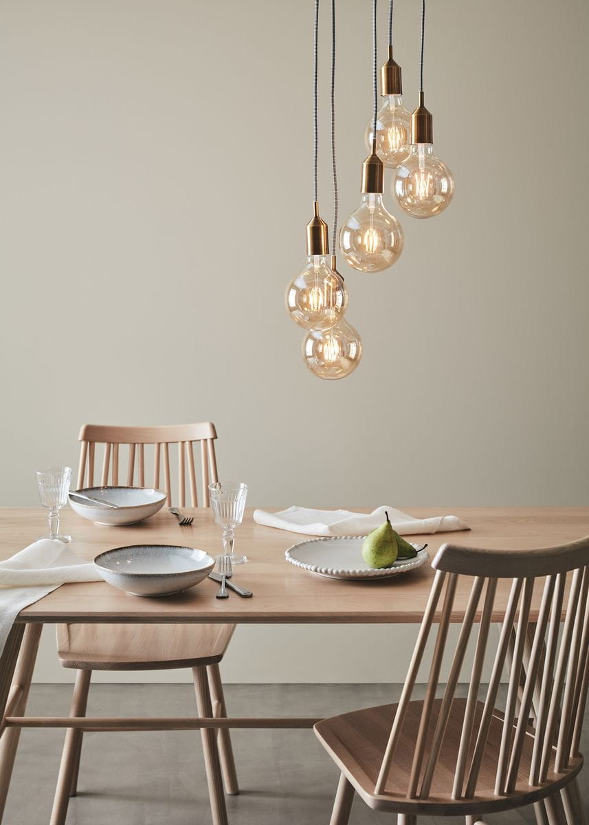 Lampa wisząca Bronx 5-punktowa w brązowej oprawie, z przezroczystymi żarówkami, zawieszona nad drewnianym stołem. W tle krzesła oraz zastawa stołowa w odcieniach szarości i bieli, na stole owoc i tekstylia.