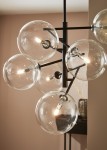 Czarna lampa wisząca Andrew z sześcioma przezroczystymi kloszami w formie kul. Klosze zawierają widoczne żarówki, umieszczone na metalowych ramionach. Produkt wisi z góry, na tle jasnej ściany, z elementami dekoracyjnymi w tle.