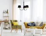 Czarna lampa wisząca Sasto 3-punktowa K-4335 z metalowymi kloszami, zawieszona w jasnym wnętrzu. W tle widać zielony fotel z poduszką, żółtą sofę oraz stół kawowy z owocami. Ściana z dużymi oknami, zasłony w neutralnym kolorze.