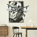 Lampa wisząca Essa 5-punktowa w czarnym kolorze z transparentnymi kloszami. Ułożona w nowoczesny sposób, z pięcioma okrągłymi kloszami, zawieszona nad drewnianym stołem w jasnym wnętrzu. Tło z grafiką oraz czarna komoda z wiklinowym akcentem.