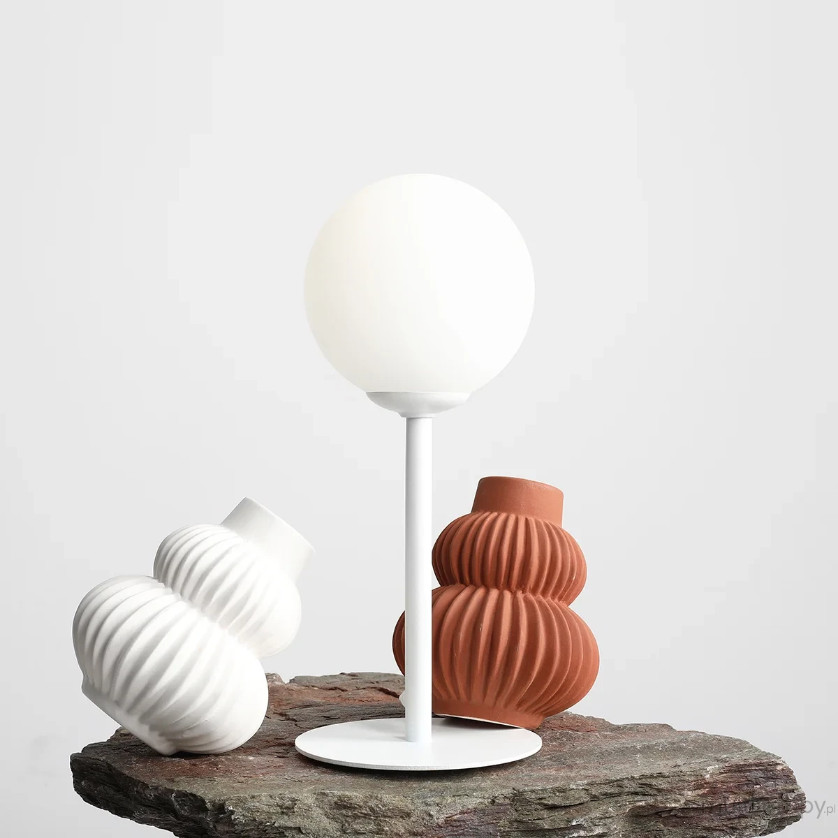 Lampa stołowa Pinne 1-punktowa, biała, z okrągłym kloszem z białego szkła na cienkiej, białej nodze. W tle widoczne dwa ceramiczne wazoniki w odcieniach bieli i terakoty, na szarej powierzchni.