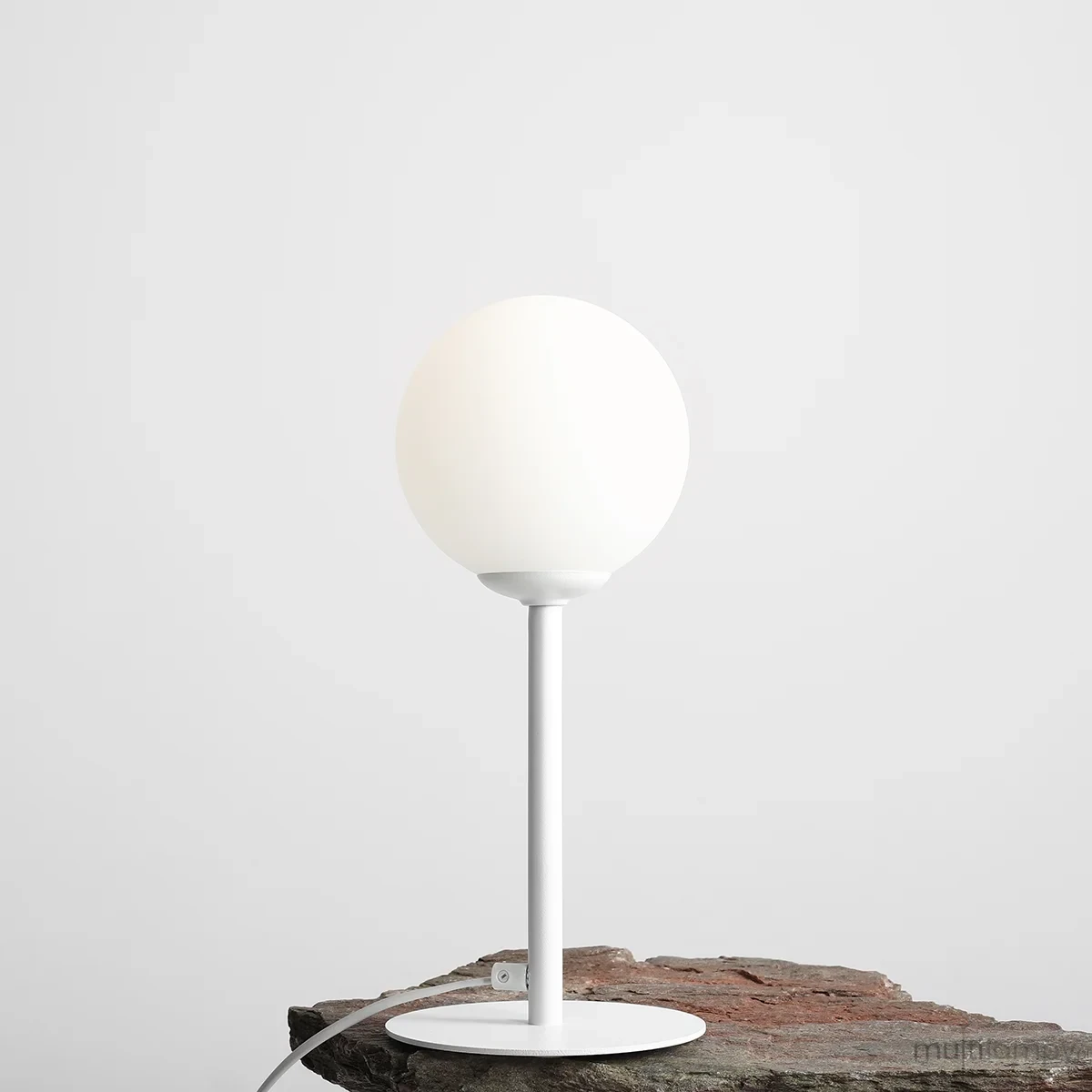Biała lampa stołowa Pinne z okrągłym kloszem, umieszczona na okrągłej podstawie. Lampa ma smukły metalowy stelaż i jest podłączona do zasilania. Ujęcie z boku na tle szarej ściany i kamiennej powierzchni.