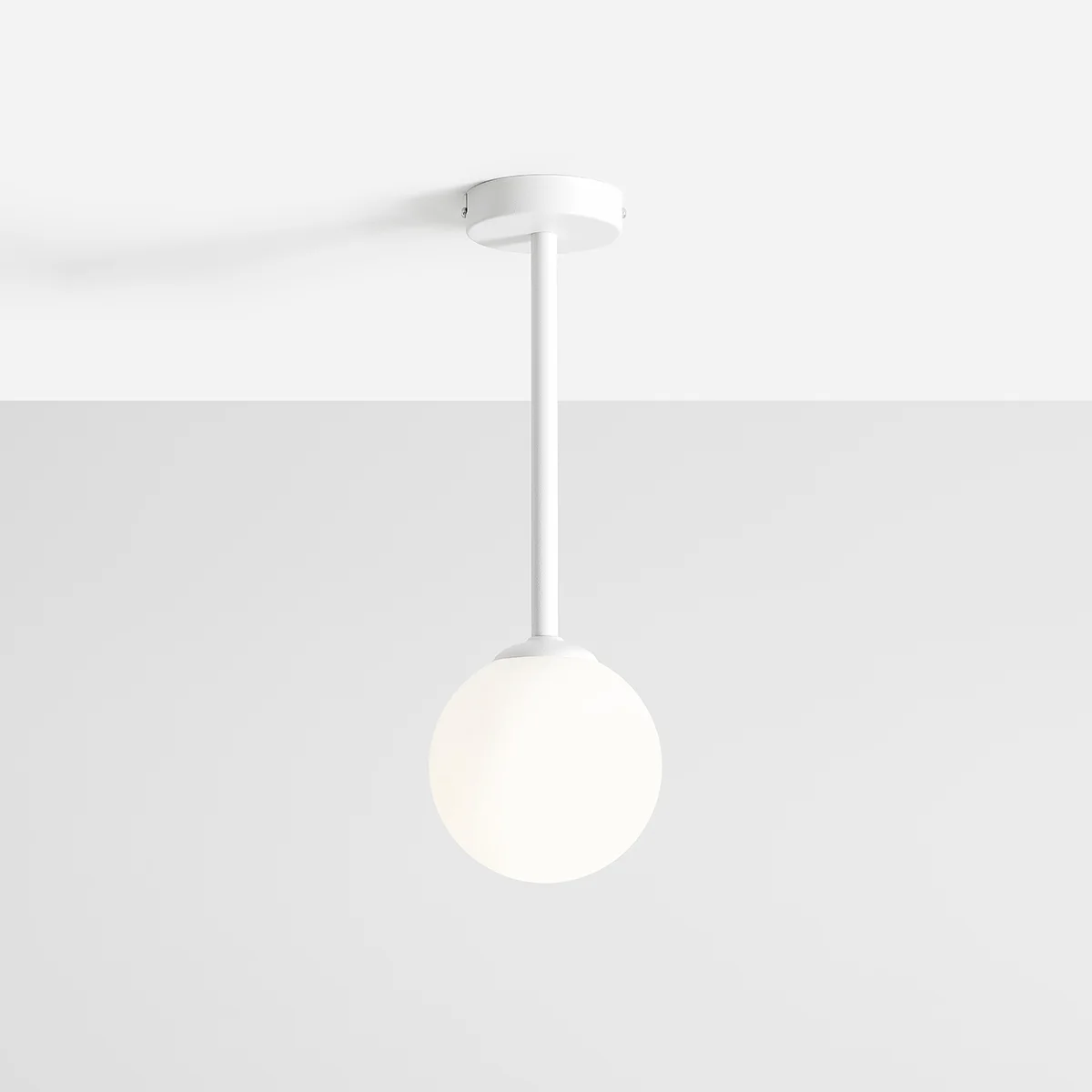 Biała lampa wisząca Pinne Short z okrągłym kloszem, zawieszona na cienkim białym pręcie. Klosz ma matową, półprzezroczystą powierzchnię, a całość umieszczona jest na jasnym tle.