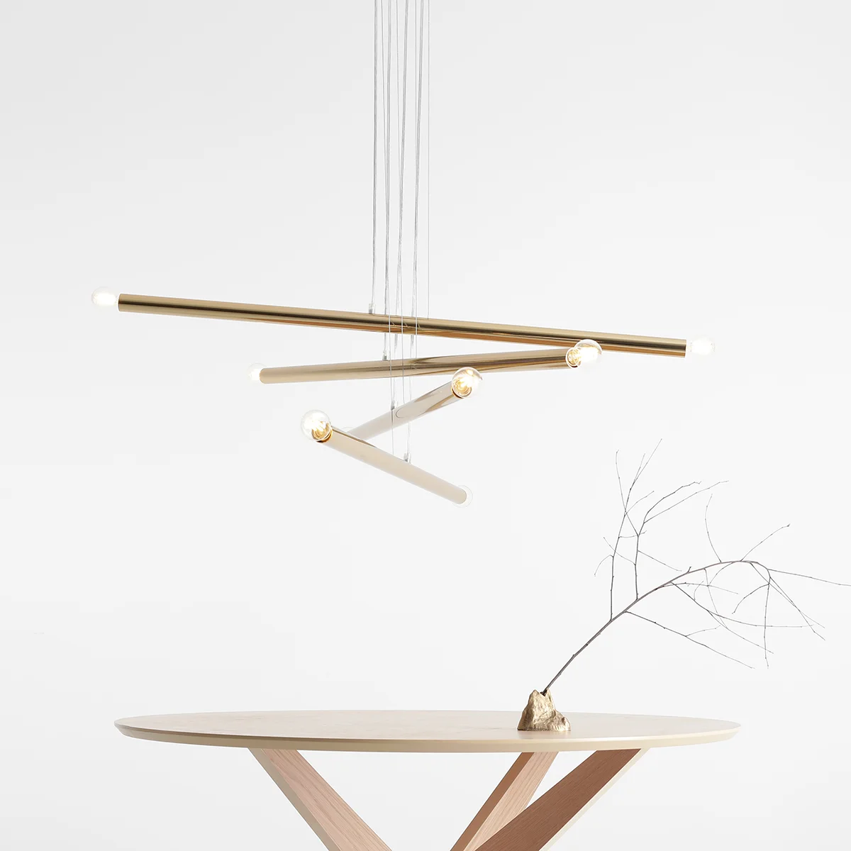 Złota lampa wisząca Tubo 8-punktowa z metalowymi prętami, zawieszona na cienkich linkach. Widoczne źródła światła na końcach prętów. Lampa umieszczona nad okrągłym stołem z jasnego drewna, w minimalistycznym wnętrzu na białym tle.