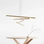Złota lampa wisząca Tubo 8-punktowa z metalowymi prętami, zawieszona na cienkich linkach. Widoczne źródła światła na końcach prętów. Lampa umieszczona nad okrągłym stołem z jasnego drewna, w minimalistycznym wnętrzu na białym tle.