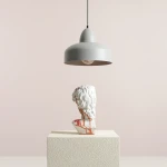 Lampa wisząca Como Gray w kolorze szarym z metalowym kloszem, zawieszona na cienkim kablu. Pod nią stoi biała figura z detalami w odcieniu brązu, na teksturowanym, jasnym podłożu. Tło w neutralnym odcieniu różu.