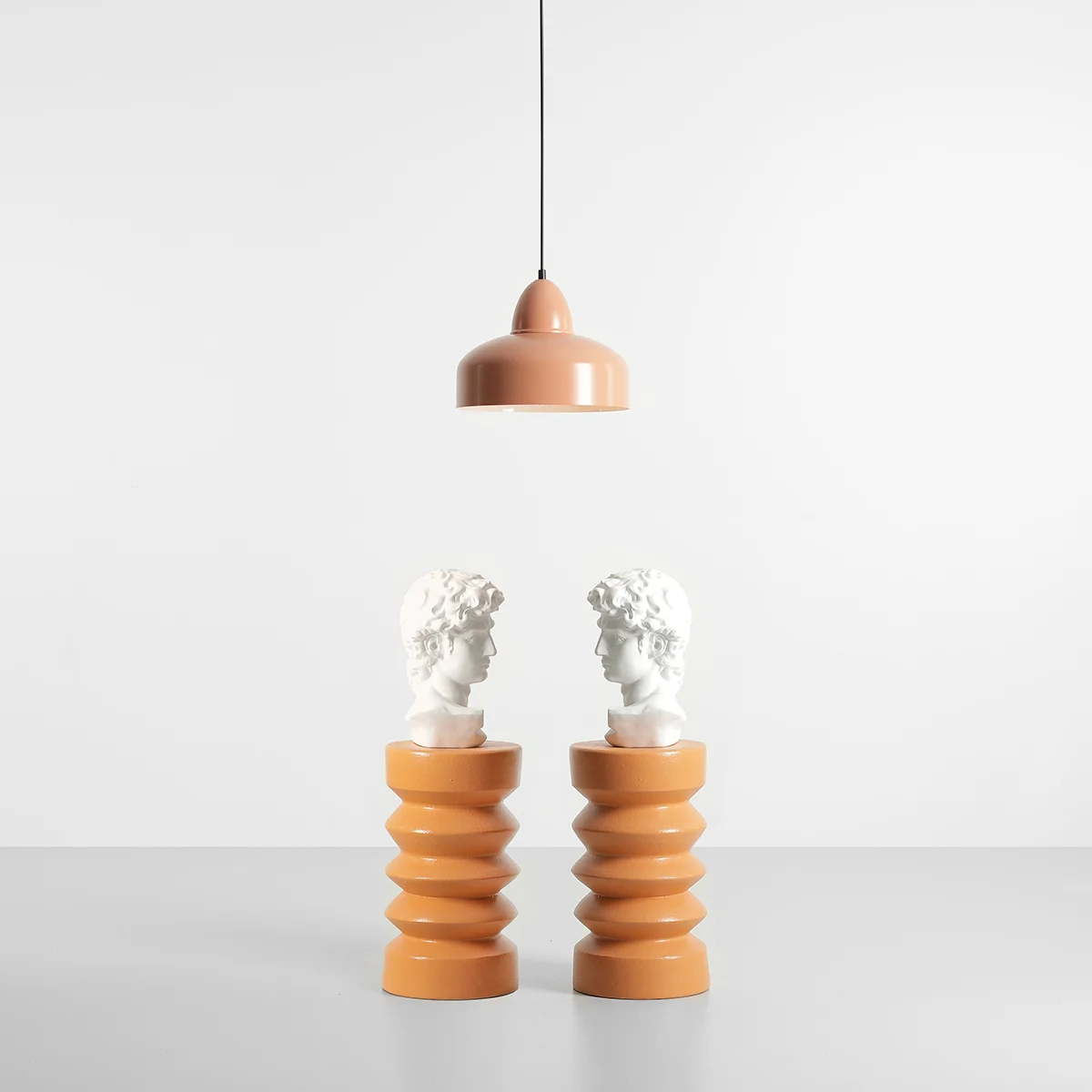 Lampa wisząca Como Coral 1-punktowa w kolorze różowym, zawieszona na czarnym przewodzie, z dwiema białymi rzeźbami głów na pomarańczowych podstawach w kształcie spirali, na jasnym tle.