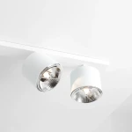 Biała lampa sufitowa 6-punktowa, z cylindrycznymi kloszami i chromowanymi wykończeniami, zamontowana na prostokątnej podstawie. Klosze skierowane są w różne kierunki, tworząc efektowne oświetlenie. Tło zdjęcia jest jasne.