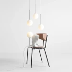 Lampa wisząca Bosso Ring 3-punktowa w białym kolorze, z trzema okrągłymi kloszami. Produkt zawieszony nad drewnianym krzesłem z czarnymi nogami, z białym kablem z włącznikiem na podłodze. Tło w jasnym kolorze.