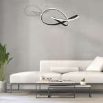 Cappio LED lampa wisząca 1-punktowa w czarnym kolorze, o nowoczesnym, abstrakcyjnym kształcie, zawieszona nad białą sofą w minimalistycznym wnętrzu. Tło w odcieniach szarości, z rośliną w doniczce oraz stolikiem kawowym z książkami i okularami.