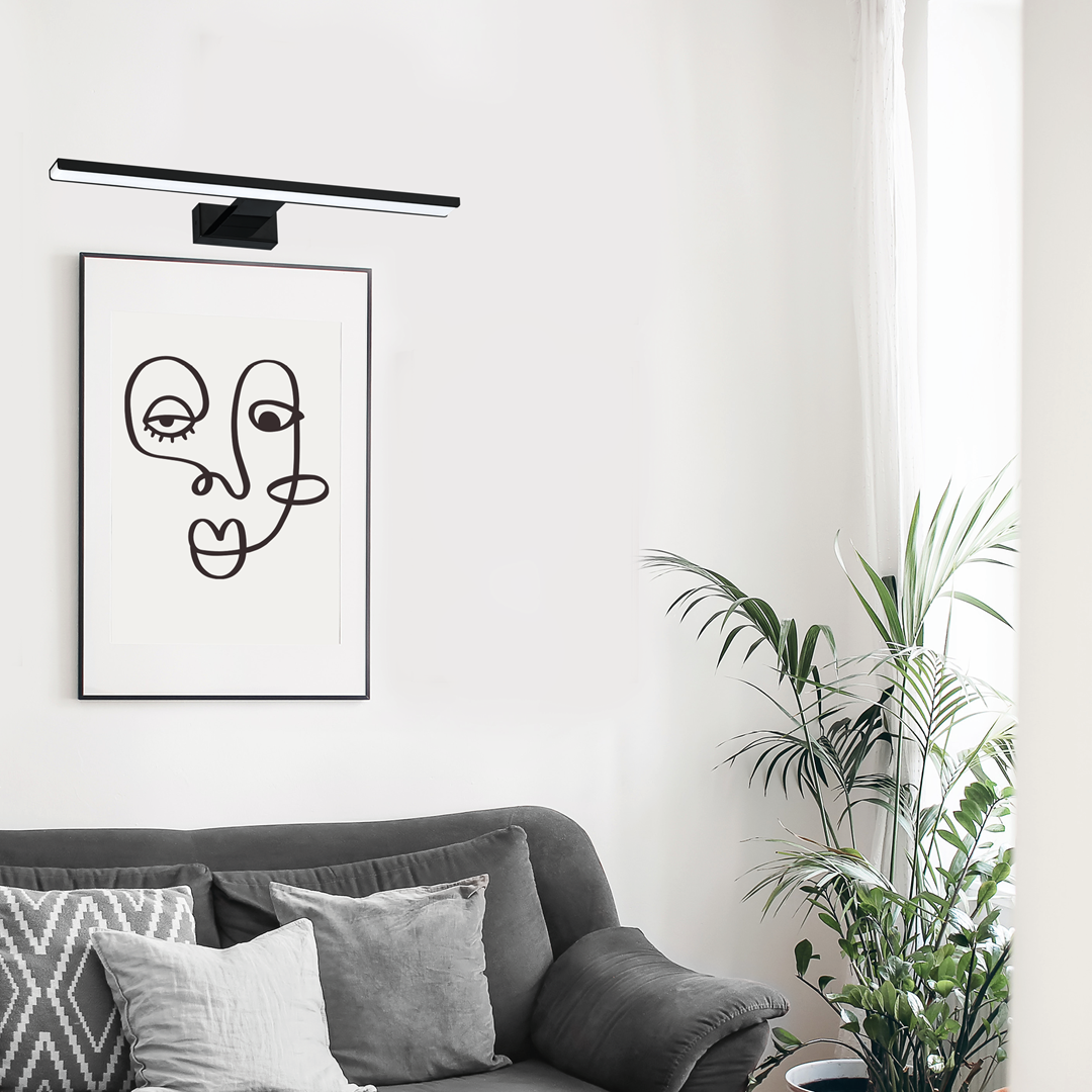 Czarny kinkiet LED Pinto 1-punktowy o długości 50 cm z zamontowanym na ścianie, skierowany w dół, nad grafiką. W tle widoczna kanapa z poduszkami oraz rośliny doniczkowe. Ściana w jasnym kolorze, minimalistyczne otoczenie.