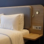 Flexy Wall LED kinkiet 1-punktowy w antycznym mosiądzu, zamontowany na ścianie obok łóżka. Elastyczna ramię lampy skierowane w dół, w pobliżu poduszki na łóżku. Tło w odcieniach szarości i drewna.