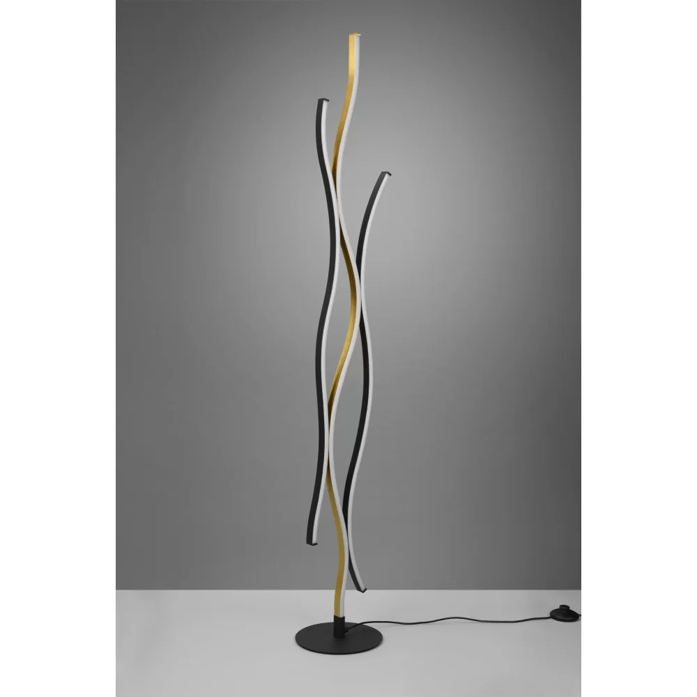 LED lampa podłogowa Blaze w kolorze mosiężnym z trzema falującymi ramionami w odcieniach czerni, bieli i złota. Stojąca na okrągłej podstawie z czarnego metalu, z kablem zasilającym na podłodze. Tło w odcieniach szarości.