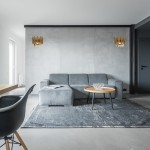 Kinkiet Madison 2-punktowy w kolorze złotym, zamontowany na szarej ścianie. Oświetlenie z geometrycznymi detalami, emituje ciepłe światło, obok szara kanapa i okrągły stół z drewnianym blatem w nowoczesnym wnętrzu.
