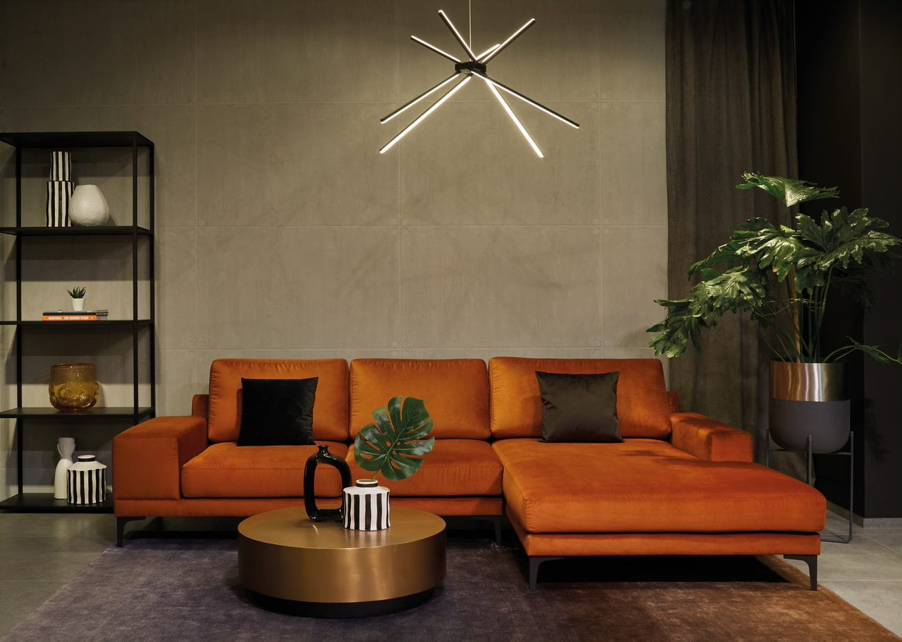 Czarna lampa wisząca Spider LED 3-punktowa, z nowoczesnym designem, zawieszona nad pomarańczową sofą. Lampa posiada trzy cienkie, prostokątne ramiona, emitujące ciepłe światło, w otoczeniu roślin i minimalistycznych mebli.