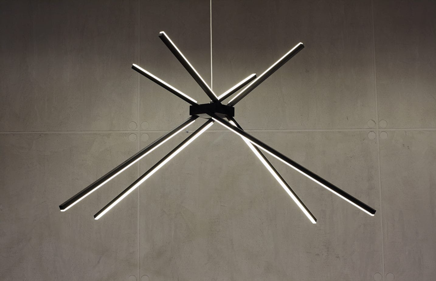 Czarna lampa wisząca Spider LED z trzema punktami świetlnymi, o nowoczesnym designie. Składa się z asymetrycznych, cienkich rur LED, umieszczonych w różnych kierunkach. Montowana na suficie, idealna do nowoczesnych wnętrz. Tło w odcieniach szarości.