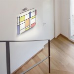Kinkiet obrazowy Barcelona LED 1-punktowy w satynowym srebrze, zamontowany na ścianie, oświetlający kolorowy obraz w stylu abstrakcyjnym. Widok z góry, na tle białej ściany i drewnianej podłogi, w jasnym wnętrzu.