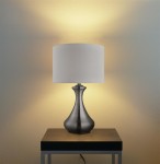 Lampa stołowa Touch z satynowego srebra, z białym abażurem, o kształcie stożkowatym, stojąca na czarnym stoliku. Świeci ciepłym światłem, tworząc przytulną atmosferę. Włączanie dotykowe, z widocznym kablem.