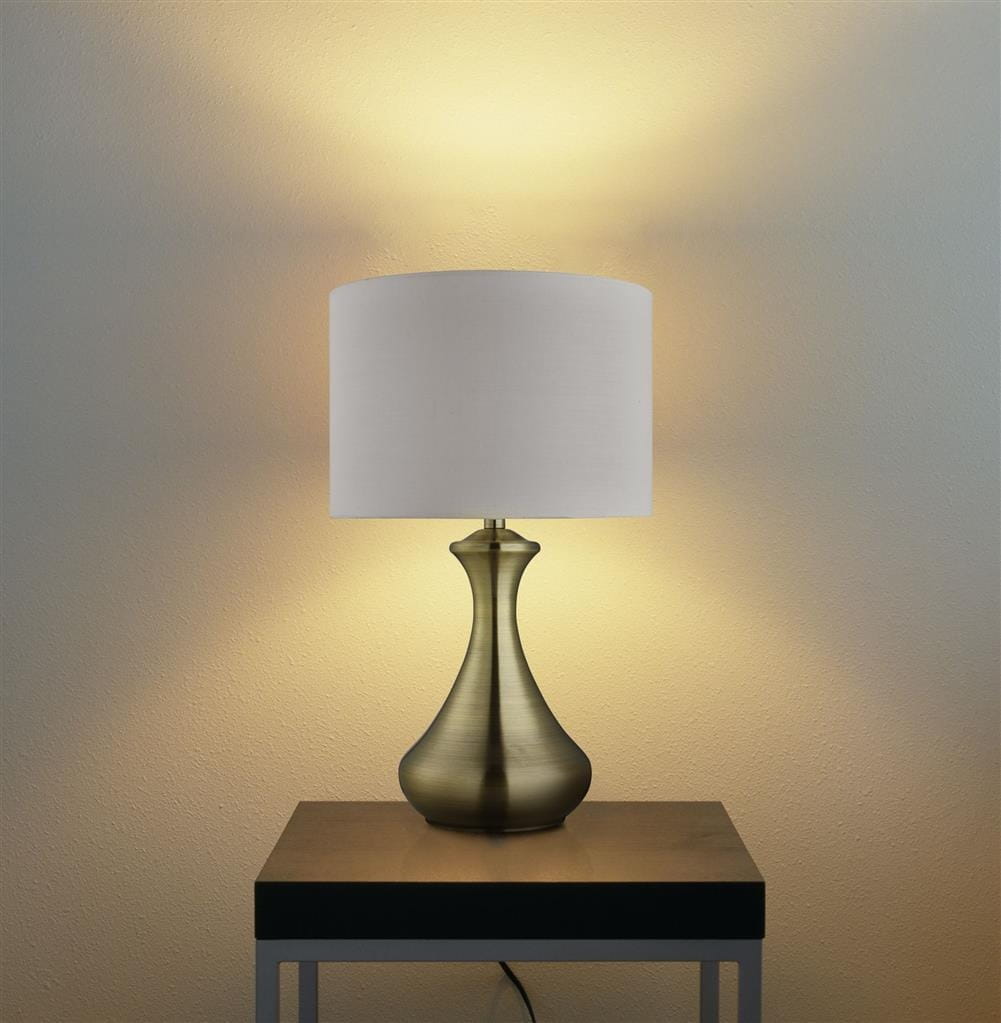 Lampa stołowa Touch 1-punktowa w antycznym mosiądzu z kremowym abażurem. Ujęcie frontalne, stojąca na czarnej podstawie, emitująca ciepłe światło na tle jasnej ściany. Elegancki kształt podstawy podkreśla nowoczesny styl.