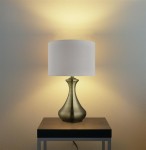 Lampa stołowa Touch 1-punktowa w antycznym mosiądzu z kremowym abażurem. Ujęcie frontalne, stojąca na czarnej podstawie, emitująca ciepłe światło na tle jasnej ściany. Elegancki kształt podstawy podkreśla nowoczesny styl.