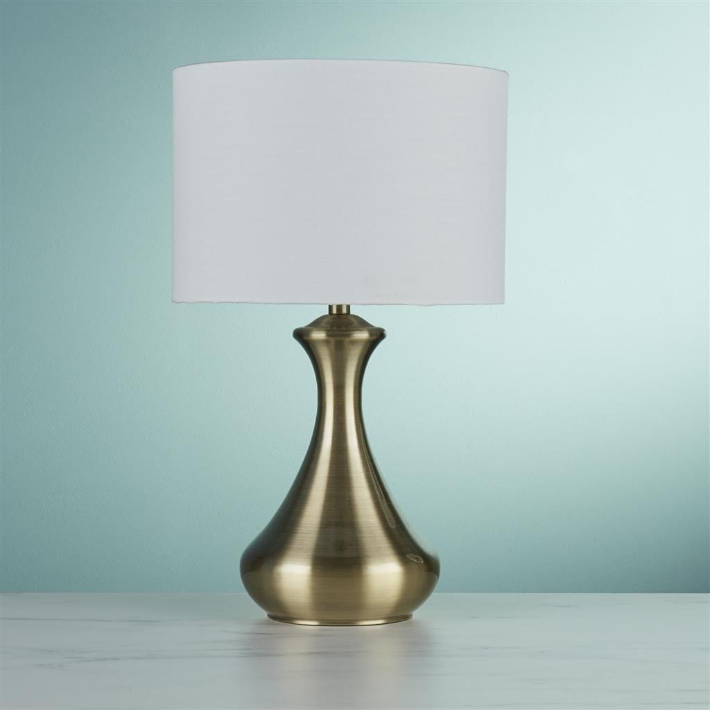 Lampa stołowa w kolorze antycznego mosiądzu z kremowym abażurem. Stojąca, o eleganckim kształcie, z zaokrągloną podstawą. Umieszczona na jasnym tle, podkreślająca nowoczesny design. Idealna do aranżacji wnętrz.