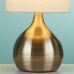 Lampa stołowa Touch z antycznym mosiądzu, o zaokrąglonym kształcie, z białym abażurem. Ujęcie z boku, na jednolitym tle. Wysoka, elegancka baza z delikatnym połyskiem, idealna do dekoracji wnętrz.
