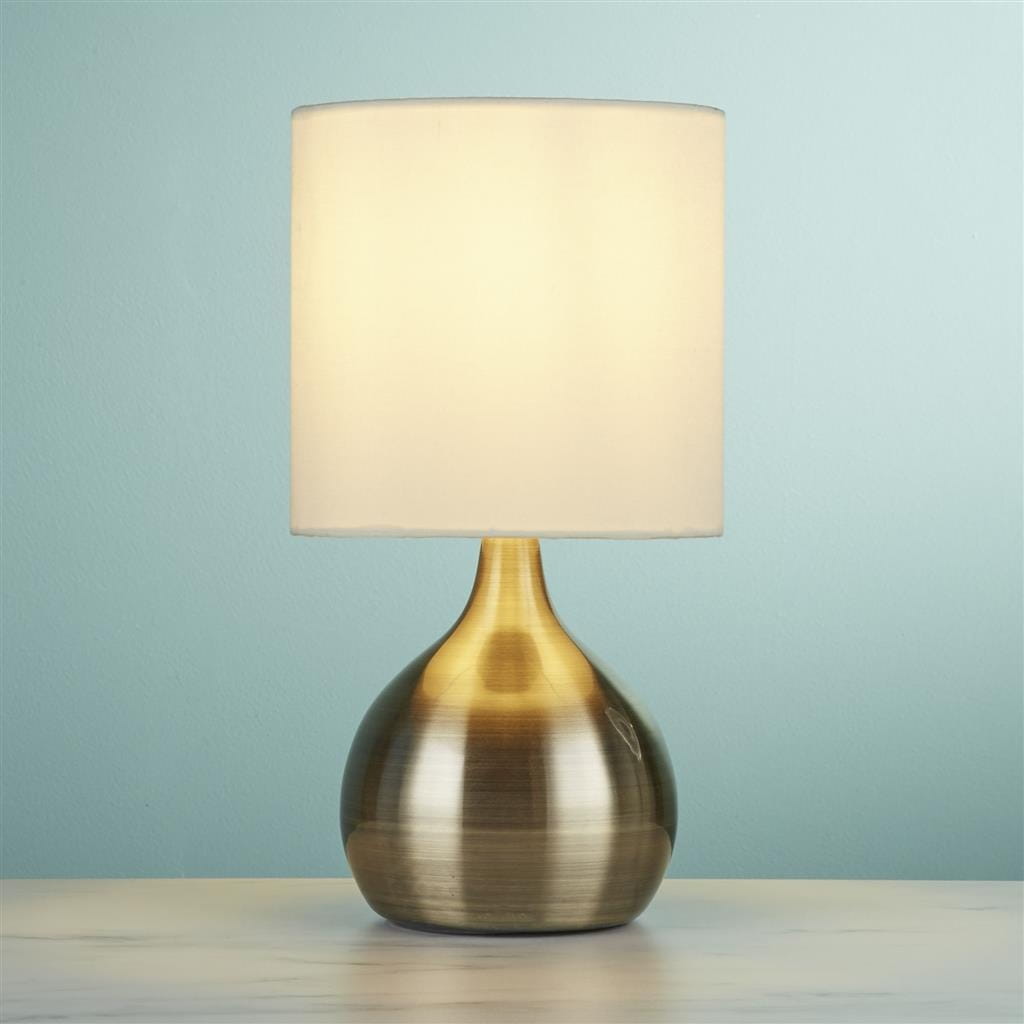 Lampa stołowa 1-punktowa w antycznym mosiądzu z białym abażurem, o zaokrąglonym kształcie podstawy. Ujęcie frontalne, lampa stoi na jasnym blacie, tło w kolorze błękitnym.
