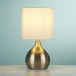 Lampa stołowa 1-punktowa w antycznym mosiądzu z białym abażurem, o zaokrąglonym kształcie podstawy. Ujęcie frontalne, lampa stoi na jasnym blacie, tło w kolorze błękitnym.