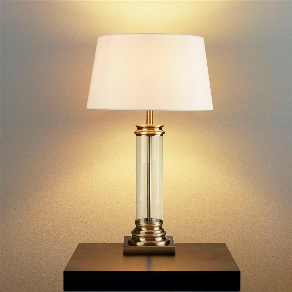 Lampa stołowa w stylu antycznym, z mosiężną podstawą i kremowym abażurem. Przezroczysta, cylindryczna część lampy z mosiężnym wykończeniem u góry. Ujęcie frontalne, lampa stoi na czarnym stole, emitując ciepłe światło na tle ściany.