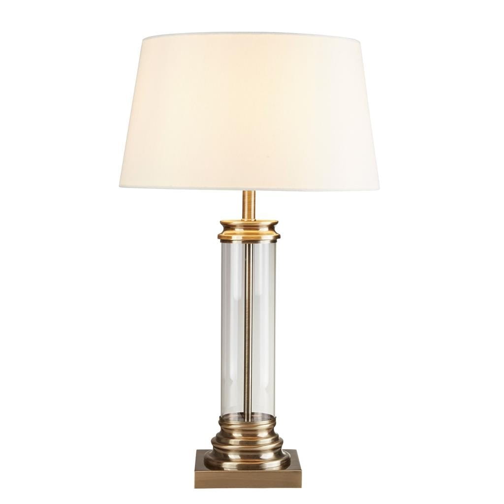 Lampa stołowa 1-punktowa z antycznym mosiądzu i kremowym kloszem. Owalny klosz na metalowej podstawie, przezroczysta kolumna z dekoracyjnymi detalami. Ujęcie frontalne, stojąca na prostokątnej podstawie.