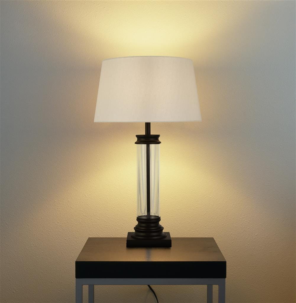 Czarna lampa stołowa z kremowym abażurem i przejrzystym, szklanym trzonem. Ujęcie frontalne, lampa stoi na czarnej podstawie na metalowym stole. Oświetlenie tworzy ciepłą atmosferę na tle jasnej ściany.