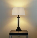 Czarna lampa stołowa z kremowym abażurem i przejrzystym, szklanym trzonem. Ujęcie frontalne, lampa stoi na czarnej podstawie na metalowym stole. Oświetlenie tworzy ciepłą atmosferę na tle jasnej ściany.