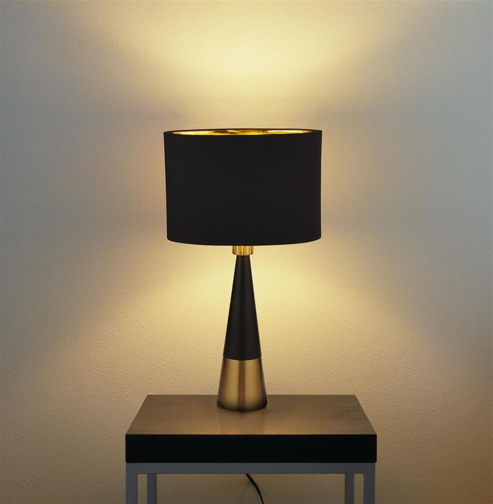 Lampa stołowa Chloe, 1-punktowa, z podstawą w antycznej miedzi i czarnym abażurem. Ustawiona na czarnym stole, emituje ciepłe światło, podkreślając elegancki design. Abażur ma złotą wewnętrzną stronę, co dodaje kontrastu.