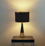 Lampa stołowa Chloe, 1-punktowa, z podstawą w antycznej miedzi i czarnym abażurem. Ustawiona na czarnym stole, emituje ciepłe światło, podkreślając elegancki design. Abażur ma złotą wewnętrzną stronę, co dodaje kontrastu.