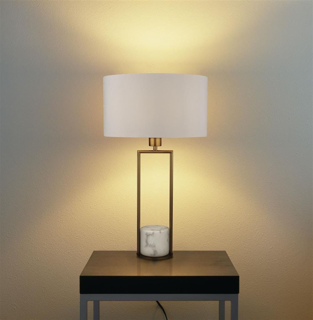 Lampa stołowa Claire 1-punktowa, złota z białym abażurem, o prostokątnym metalowym stelażu i marmurową podstawą. Ujęcie frontalne, lampa stoi na czarnym stole, w tle delikatne oświetlenie ściany.
