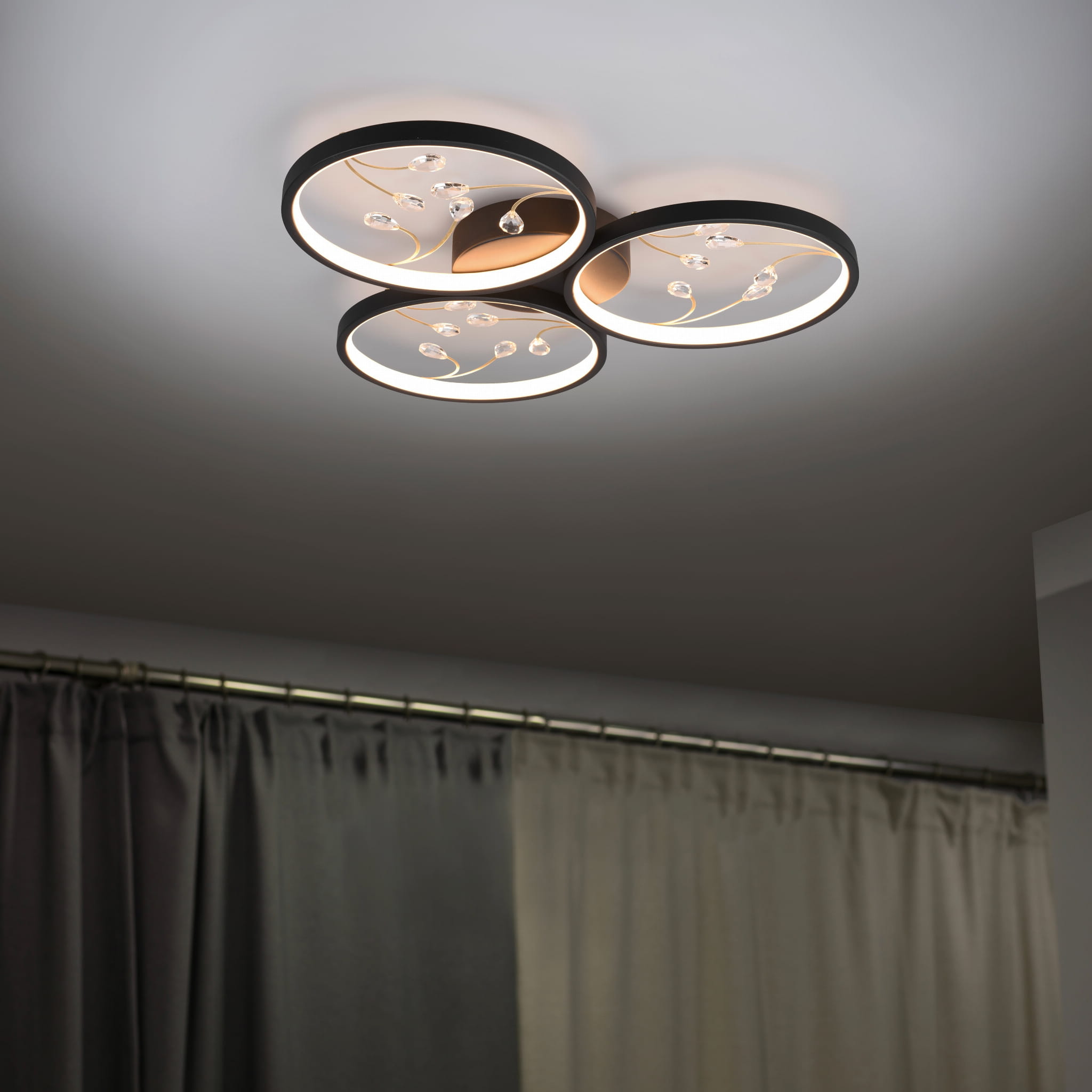 Czarna lampa sufitowa LED Groovy 3000K z trzema okrągłymi punktami świetlnymi. Widoczne dekoracyjne detale w kształcie linii oraz kryształki. Ujęcie z dołu, montowana na suficie, w tle zasłony w odcieniach szarości.