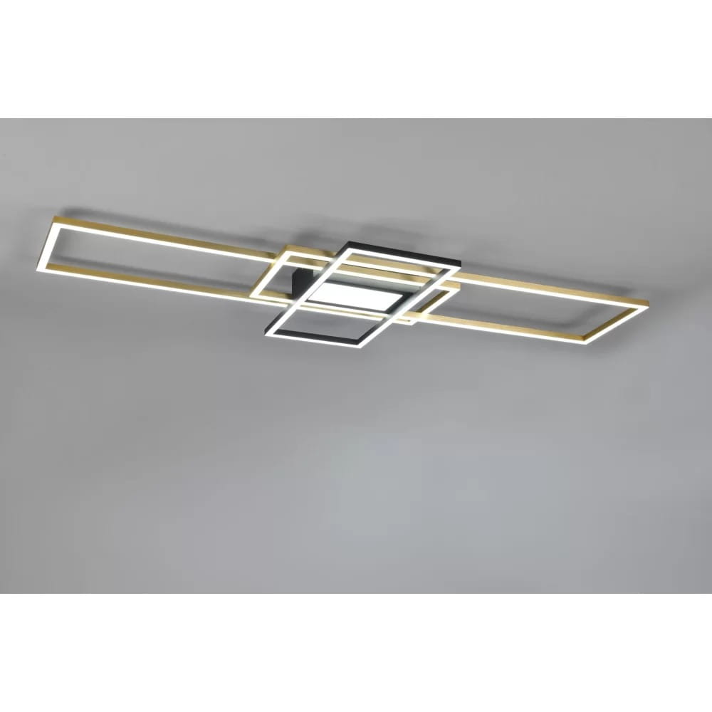 Czteropunktowa lampa sufitowa Irvine w kolorze mosiądzu i czerni. Posiada prostokątne ramy w różnych rozmiarach, z wbudowanym oświetleniem LED. Zamontowana na suficie, z widocznym pilotem do sterowania. Tło szare, minimalistyczne.