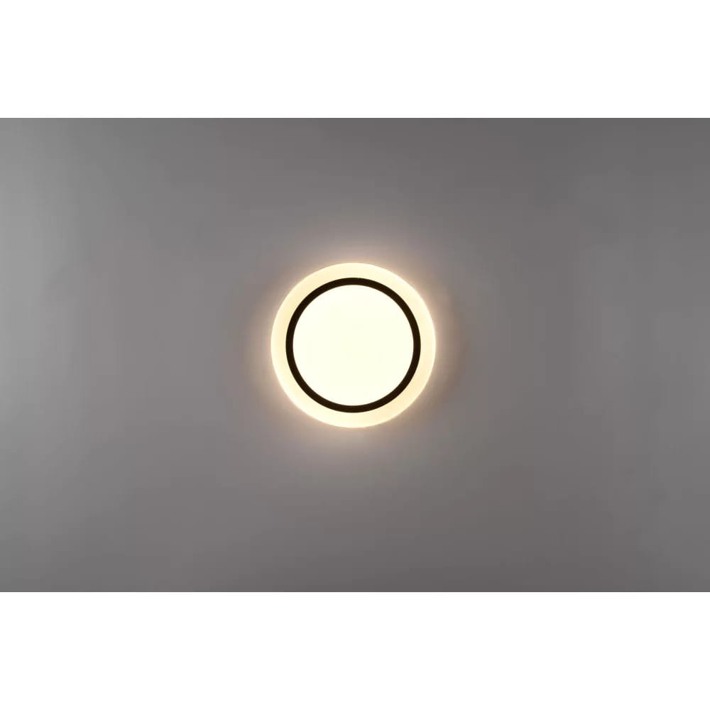 Plafon Atria D27 LED w kolorze białym z czarnym obrzeżem, okrągły, zamontowany na suficie. Emituje ciepłe światło, widoczny pilot do sterowania. Tło jasne, proste, co podkreśla design lampy.
