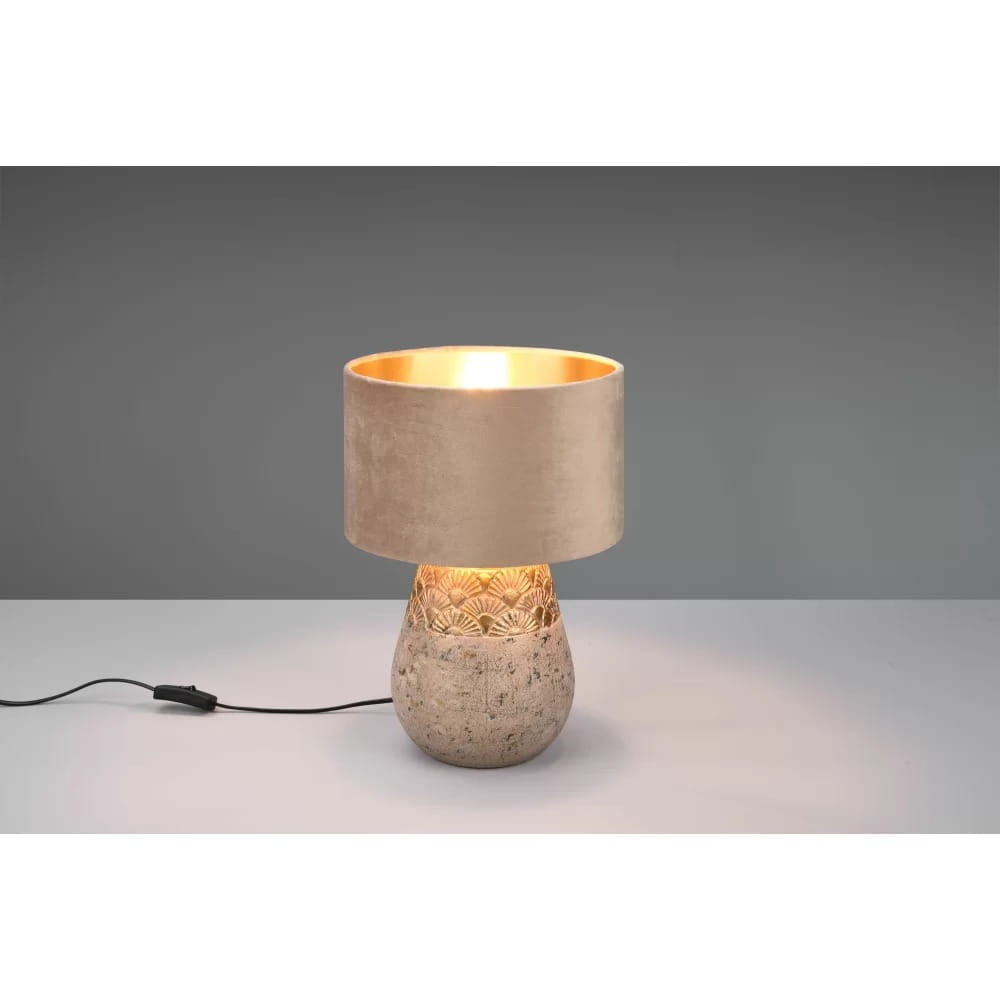 Lampa stołowa Kiran 1-punktowa w kolorze szarym i beżowym, z okrągłym kloszem z aksamitnej tkaniny i dekoracyjną podstawą z fakturowanego materiału. Ujęcie z boku, włączona, na prostym tle.