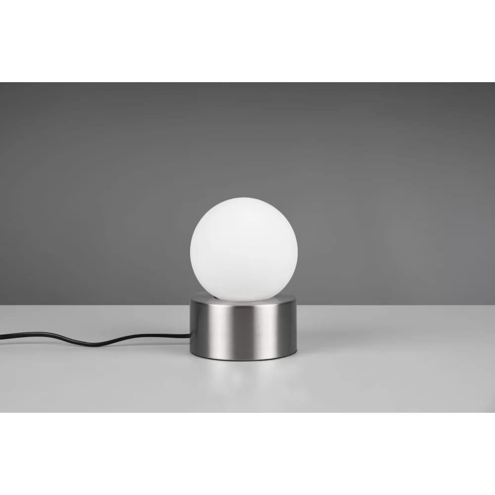 Lampa stołowa Countess w kolorze niklu z białym, okrągłym kloszem, umieszczona na metalowej podstawie. Produkt stoi na jednolitej powierzchni, z widocznym kablem zasilającym. Tło w odcieniach szarości.