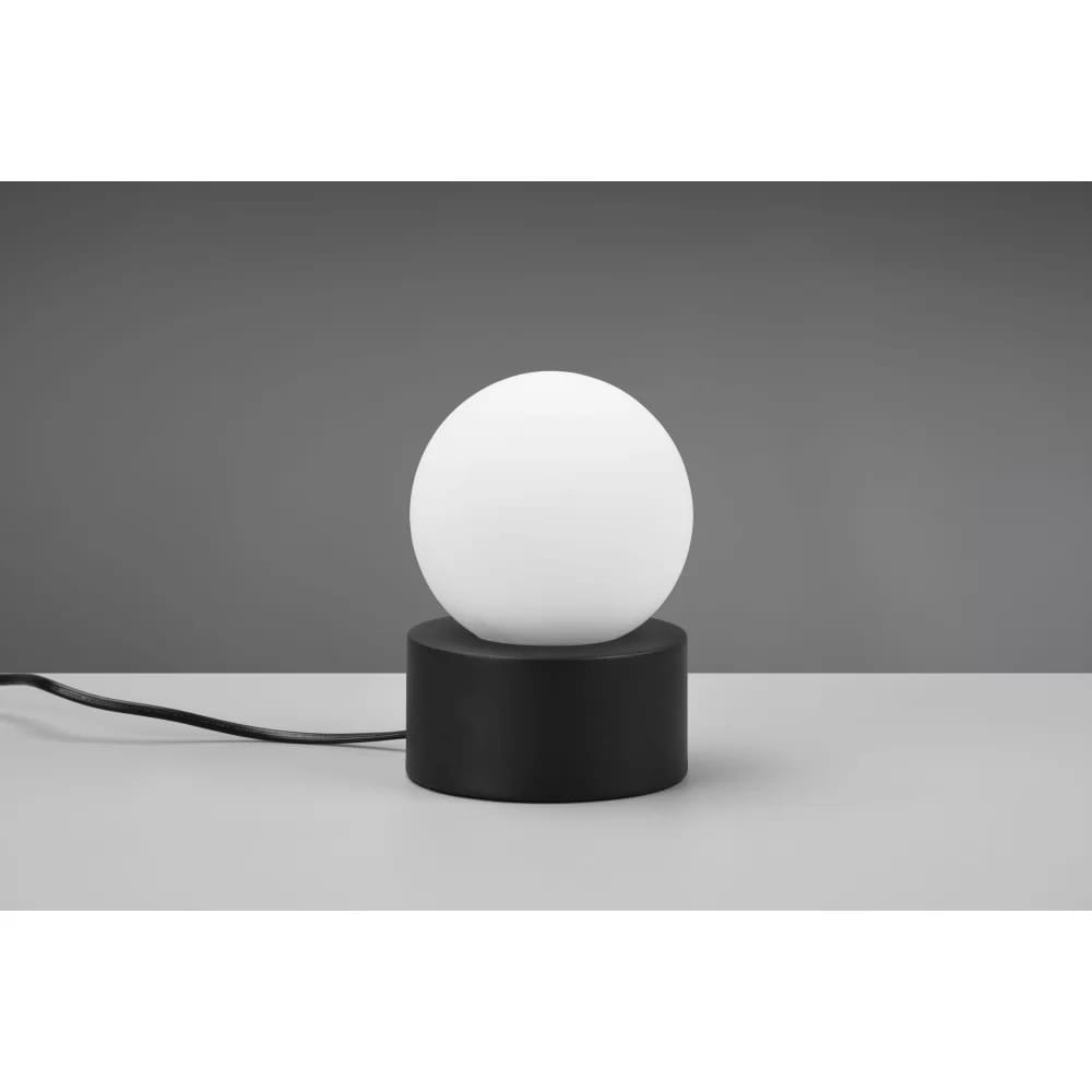 Czarna lampa stołowa Countess z białym, okrągłym kloszem, umieszczona na okrągłej podstawie. Widoczny kabel zasilający. Lampa prezentuje minimalistyczny design, idealna do nowoczesnych wnętrz. Ujęcie z boku na neutralnym tle.