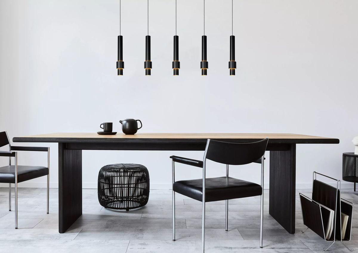 Czarna lampa wisząca Margary LED 5-punktowa, z pięcioma cylindrycznymi kloszami z metalowymi detalami, zawieszona nad drewnianym stołem. W tle widać minimalistyczne krzesła i dekoracyjne elementy. Proste, jasne tło podkreśla nowoczesny design.
