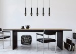 Czarna lampa wisząca Margary LED 5-punktowa, z pięcioma cylindrycznymi kloszami z metalowymi detalami, zawieszona nad drewnianym stołem. W tle widać minimalistyczne krzesła i dekoracyjne elementy. Proste, jasne tło podkreśla nowoczesny design.