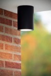 Czarna lampa sufitowa elewacyjna Volantis 1-punktowa, w formie cylindrycznej, zamontowana pod sufitem. Widoczna okrągła część emitująca światło, otoczona gładką, matową powierzchnią. Tło z ceglanym murem oraz rozmytym zielonym otoczeniem.