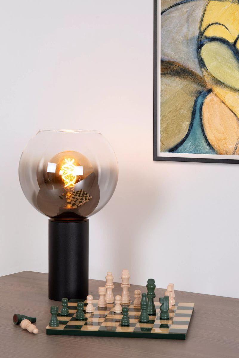 Lampa stołowa Marius 1-punktowa w czarnej podstawie z przezroczystą, szklaną kopułą. Widoczna żarówka o spiralnym kształcie. Na stole szachy w kolorze jasnym i ciemnym. Tło w jasnym kolorze, obok obraz w ramie.