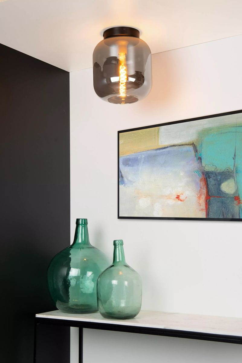 Czarna lampa sufitowa Joanet 1-punktowa z przezroczystym kloszem, zamontowana na suficie. Widoczna żarówka w kształcie spirali. Tło w neutralnych kolorach, z dekoracjami w postaci zielonych butelek i obrazu.